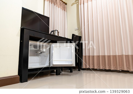 Low angle view of the opened mini fridge with water bottles placed under wooden table near the curtains in a hotel room. Low angle view of the opened mini fridge with water bottles placed under wooden table near the curtains in a hotel room. 139329063
