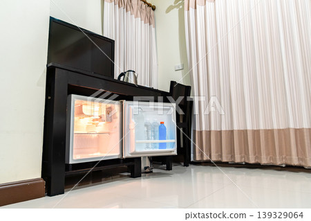 Low angle view of the opened mini fridge with water bottles placed under wooden table near the curtains in a hotel room. Low angle view of the opened mini fridge with water bottles placed under wooden table near the curtains in a hotel room. 139329064