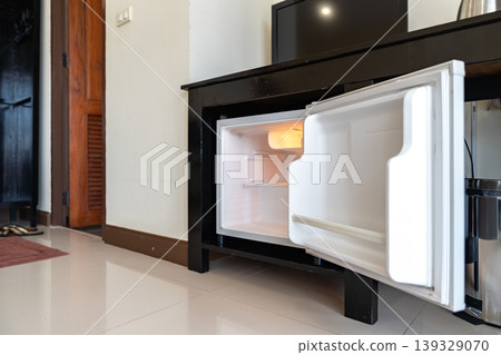 Low angle view of the opened mini fridge with water bottles placed under wooden table near the curtains in a hotel room. Low angle view of the opened mini fridge with water bottles placed under wooden table near the curtains in a hotel room. 139329070