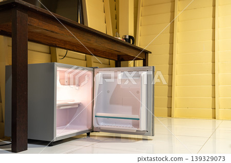 Low angle view of the opened mini fridge with water bottles placed under wooden table near the curtains in a hotel room. Low angle view of the opened mini fridge with water bottles placed under wooden table near the curtains in a hotel room. 139329073