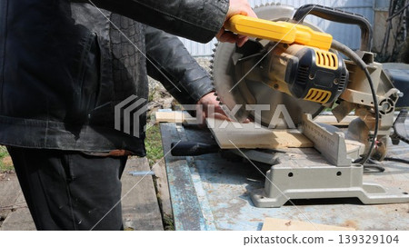 A close-up of a workman working on a miter saw during the process of cutting a board, the hands of a carpenter on the lever of a circular saw, cutting the end of a board A close-up of a workman working on a miter saw during the process of cutting a board, the hands of a carpenter on the lever of a circular saw, cutting the end of a board 139329104