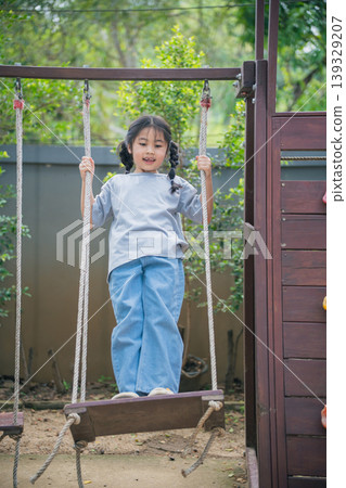 Young girl on swing enjoying outdoor playtime in nature, wearing casual clothing, smiling joyfully in a playground setting surrounded by greenery Young girl on swing enjoying outdoor playtime in nature, wearing casual clothing, smiling joyfully in a playground setting surrounded by greenery 139329207