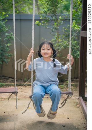 Happy young girl sitting on a wooden swing in a serene outdoor playground surrounded by greenery, enjoying a sunny day with a joyful expression Happy young girl sitting on a wooden swing in a serene outdoor playground surrounded by greenery, enjoying a sunny day with a joyful expression 139329210