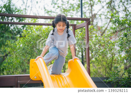 Happy child playing on yellow slide in outdoor playground, enjoying sunny day with trees and greenery in background, carefree childhood fun and laughter 139329211