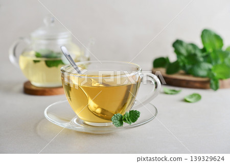 Glass cup of fresh mint tea on a light background with a glass teapot and mint leaves on a wooden board 139329624