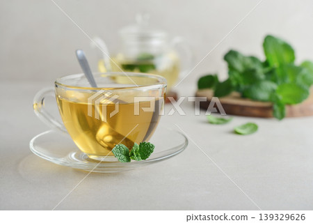 Glass cup of fresh mint tea on a light background with a glass teapot and mint leaves on a wooden board 139329626