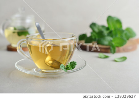 Glass cup of fresh mint tea on a light background with a glass teapot and mint leaves on a wooden board 139329627