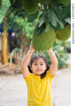 Young Child Joyfully Reaching for Large Jackfruit on Tree in Lush Green Garden Setting in Bright Summer Daylight 139329803