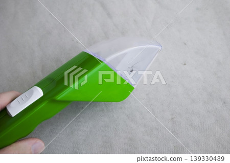 A close up of a hand holding a green insect and spider catcher with a spider inside, against textured fabric. A close up of a hand holding a green insect and spider catcher with a spider inside, against textured fabric. 139330489