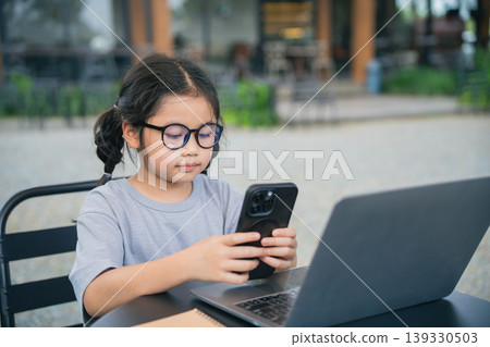 Young girl using smartphone while sitting at outdoor cafe table with laptop, focused and engaged in digital communication and learning activities 139330503