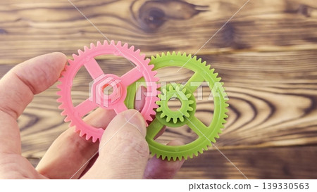 Hand holding pink and green plastic gears, showcasing their interlocking teeth and design, against a wooden background emphasizing mechanical connection and functionality Hand holding pink and green plastic gears, showcasing their interlocking teeth and design, against a wooden background emphasizing mechanical connection and functionality 139330563