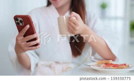 Asian women using smartphone drinking coffee eating breakfast toast jam morning routine at home relaxing happy lifestyle browsing mobile phone technology 139330775