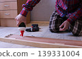 A man in a plaid suit sits on the floor in a bedroom and applies glue from a tube to the inside of a wooden bed's side plank, DIY home furniture repair 139331004