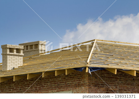 Close-up detail of wooden roof of new brick house with two white chimneys under construction. Timber frame of natural materials against bright sky. Professional building and reconstruction concept. Close-up detail of wooden roof of new brick house with two white chimneys under construction. Timber frame of natural materials against bright sky. Professional building and reconstruction concept. 139331371