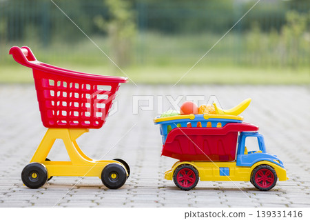 Bright plastic colorful toys for children outdoors on sunny summer day. Car truck carrying basket with toy fruit and vegetables and shopping pushcart. Bright plastic colorful toys for children outdoors on sunny summer day. Car truck carrying basket with toy fruit and vegetables and shopping pushcart. 139331416