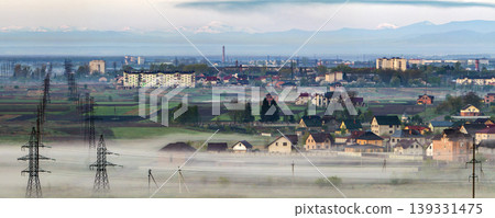 Beautiful panorama of long electric high voltage power line rows stretching to horizon through spring fields and trees under dense morning fog on distant Carpathian mountains range background. Beautiful panorama of long electric high voltage power line rows stretching to horizon through spring fields and trees under dense morning fog on distant Carpathian mountains range background. 139331475
