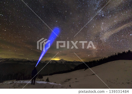 Back view of man with head flashlight standing on snowy valley under beautiful dark blue winter starry sky, bright blue beam on stars and mountains ridge background. Night photography, light painting. Back view of man with head flashlight standing on snowy valley under beautiful dark blue winter starry sky, bright blue beam on stars and mountains ridge background. Night photography, light painting. 139331484