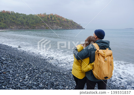 Couple enjoys Turkish coastline. Sitting on stones, watching sea waves, relaxing together, slow romantic moment near calm blue water under soft winter light. 139331837