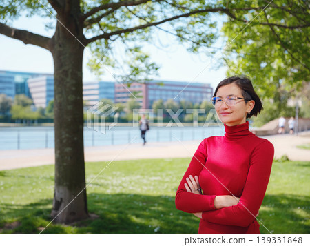 Woman in glasses and red turtleneck, dressed in business style, stands in center of Frankfurt, surrounded by modern buildings and urban life, embodying professionalism and confidence 139331848