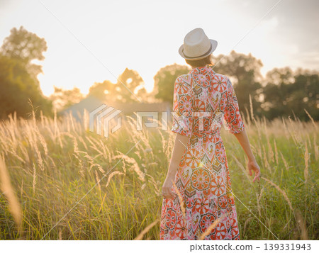 Young woman walking through picturesque European field in late summer. Golden sunlight, lush greenery, and serene rural atmosphere create peaceful countryside scene. Young woman walking through picturesque European field in late summer. Golden sunlight, lush greenery, and serene rural atmosphere create peaceful countryside scene. 139331943