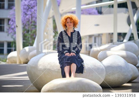 Woman in black dress sitting on stone surface adjusting clothing Woman in black dress sitting on stone surface adjusting clothing 139331958