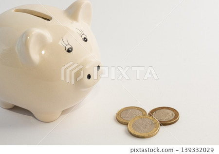 Piggy bank in the shape of a pig with three 1 euro coins next to it on a white background. Piggy bank in the shape of a pig with three 1 euro coins next to it on a white background. 139332029