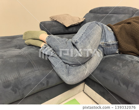 A man curled up on a blue corner sofa in jeans and socks, a cozy home interior for relaxation and a casual lifestyle A man curled up on a blue corner sofa in jeans and socks, a cozy home interior for relaxation and a casual lifestyle 139332076