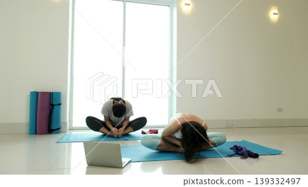 Two women performing seated forward fold yoga pose on mats during home workout, enhancing flexibility, hip opening and muscle recovery, promoting relaxation, mindfulness and healthy lifestyle in Two women performing seated forward fold yoga pose on mats during home workout, enhancing flexibility, hip opening and muscle recovery, promoting relaxation, mindfulness and healthy lifestyle in 139332497