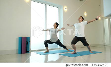 Man and woman practicing warrior yoga pose with open arms at home, improving balance, strength and flexibility, enhancing posture and full body coordination in bright minimalist indoor fitness space Man and woman practicing warrior yoga pose with open arms at home, improving balance, strength and flexibility, enhancing posture and full body coordination in bright minimalist indoor fitness space 139332528