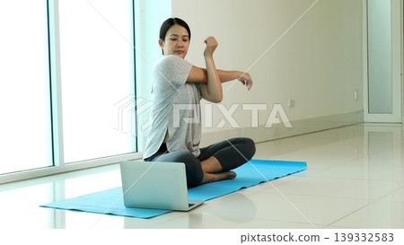 Woman stretching arm during online yoga class at home using laptop, sitting on mat and warming up body, highlighting fitness routine, healthy lifestyle, flexibility training and digital wellness Woman stretching arm during online yoga class at home using laptop, sitting on mat and warming up body, highlighting fitness routine, healthy lifestyle, flexibility training and digital wellness 139332583