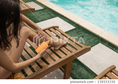 Woman applying sunscreen lotion to leg by sunny pool on a wooden lounge chair Woman applying sunscreen lotion to leg by sunny pool on a wooden lounge chair 139333150