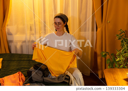 Caucasian young woman organizing clothes in a bedroom filled with sunset light. She folds laundry carefully. Clean home and daily routine theme. Mid shot Caucasian young woman organizing clothes in a bedroom filled with sunset light. She folds laundry carefully. Clean home and daily routine theme. Mid shot 139333304