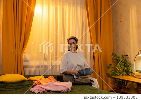 Young Caucasian woman folds clothes in her room during warm sunset light. She organizes garments indoors. Home cleaning and space organization concept. 139333305
