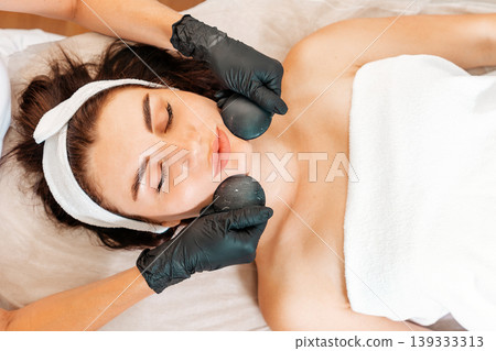 Beautician in gloves gives a facial massage with stones to the client in salon. Close up of female face. Top view. Concept of cosmetology and beauty procedures 139333313