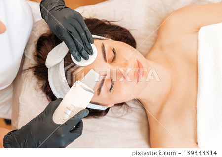 Cleaning procedure at the beauty salon. Cosmetologist cleans the client's forehead using a ultrasonic device. Close up. Top view. Concept of cosmetology and beauty 139333314