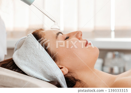 Darsonval cosmetology apparatus. Beautician cleans the client's forehead using a electric device. Close up. Professional rejuvenation procedure in salon Darsonval cosmetology apparatus. Beautician cleans the client's forehead using a electric device. Close up. Professional rejuvenation procedure in salon 139333315