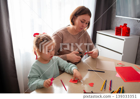 Mid shot of young Caucasian woman teaches how to carve hearts with a cute little girl while sitting at a table. Preparing for Mother's Day and Valentine's Day. The concept of preschool education Mid shot of young Caucasian woman teaches how to carve hearts with a cute little girl while sitting at a table. Preparing for Mother's Day and Valentine's Day. The concept of preschool education 139333351