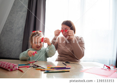 Funny mother teaches her little daughter how to cut hearts out of paper. The family is sitting at a table in the living room and making holiday crafts. The concept of Mother's Day 139333353