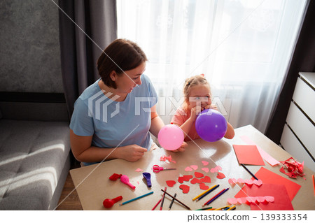 Top view of young Caucasian mother and her little daughter inflate balloons and make decorations for the holiday. Family sits at the table and makes festive decorations. Concept of Mother's Day Top view of young Caucasian mother and her little daughter inflate balloons and make decorations for the holiday. Family sits at the table and makes festive decorations. Concept of Mother's Day 139333354
