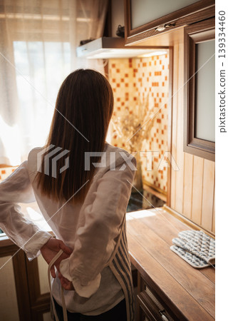 Back shot of a girl putting on an apron in kitchen before cooking. Preparations before cooking, put on an apron Back shot of a girl putting on an apron in kitchen before cooking. Preparations before cooking, put on an apron 139334460