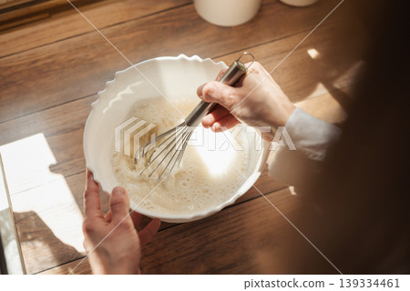 Girl whipping dough for baking bread. Close up of a wife cooking in kitchen, using whisk to stir different ingredients for liquid dough Girl whipping dough for baking bread. Close up of a wife cooking in kitchen, using whisk to stir different ingredients for liquid dough 139334461