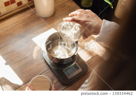 Measuring flour with electronic kitchen scales for cooking something. Girl pouring flour to the metal cup for precise measuring Measuring flour with electronic kitchen scales for cooking something. Girl pouring flour to the metal cup for precise measuring 139334462