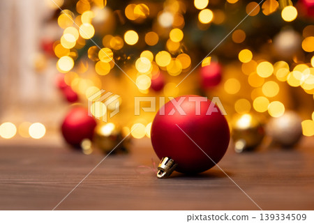 Close up on Christmas tree ball decoration on wooden floor with background of bright Christmas tree. Celebrating Christmas, holiday background, festive winter mood Close up on Christmas tree ball decoration on wooden floor with background of bright Christmas tree. Celebrating Christmas, holiday background, festive winter mood 139334509