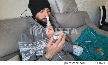 Bearded man using a medical nebulizer for respiratory therapy on a sofa, domestic health treatment, wellness routine, illness recovery Bearded man using a medical nebulizer for respiratory therapy on a sofa, domestic health treatment, wellness routine, illness recovery 139334671