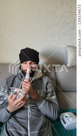 Bearded man using a medical nebulizer for respiratory therapy on a sofa, domestic health treatment, wellness routine, illness recovery Bearded man using a medical nebulizer for respiratory therapy on a sofa, domestic health treatment, wellness routine, illness recovery 139334673