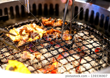 Close-up of delicious Korean barbecue meat cooking over glowing charcoal on a metal grill. Metal chopsticks picking up a piece of grilled pork. Authentic Asian cuisine and restaurant dining. Close-up of delicious Korean barbecue meat cooking over glowing charcoal on a metal grill. Metal chopsticks picking up a piece of grilled pork. Authentic Asian cuisine and restaurant dining. 139336907
