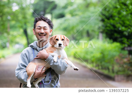 A man's smiling face as he holds his beloved beagle against a backdrop of fresh greenery, a close-up shot of him looking directly at the camera during a happy stroll in the park. A man's smiling face as he holds his beloved beagle against a backdrop of fresh greenery, a close-up shot of him looking directly at the camera during a happy stroll in the park. 139337225
