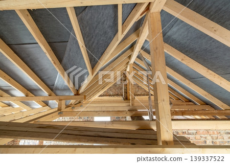 Attic space of a building under construction with wooden beams of a roof structure and brick walls. Real estate development concept. Attic space of a building under construction with wooden beams of a roof structure and brick walls. Real estate development concept. 139337522