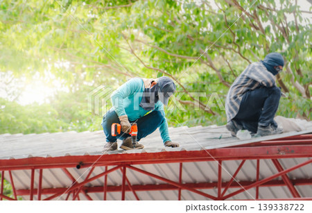 Roofer working in special protective work wear gloves, using air or pneumatic nail gun installing asphalt shingle on top of the new roof, Concept of residential building under construction. Roofer working in special protective work wear gloves, using air or pneumatic nail gun installing asphalt shingle on top of the new roof, Concept of residential building under construction. 139338722