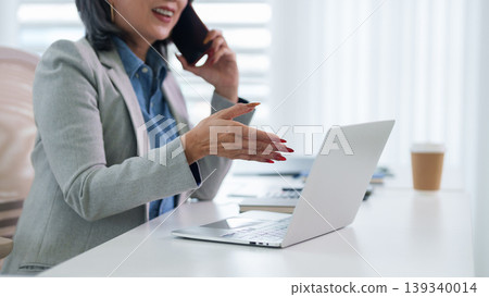 Asian businesswoman talking phone call working laptop computer at bright modern office desk professional adult women multitasking mobile conversation using Asian businesswoman talking phone call working laptop computer at bright modern office desk professional adult women multitasking mobile conversation using 139340014